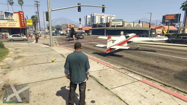 A light aircraft standing on a city boulevard beside the traffic lights, shopfronts and mountains behind it.